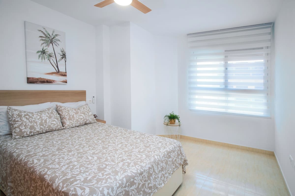 A bedroom with a bed, a ceiling fan, a window with blinds, and a wall-mounted plant.