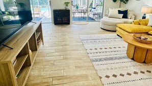 A spacious living room with a large TV, wooden flooring, a patterned rug, and a comfortable seating area with a sofa and yellow cushions.
