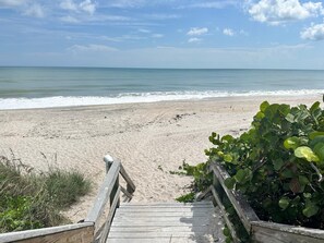 A wooden boardwalk leading to a sandy beach with calm waters and a clear sky.