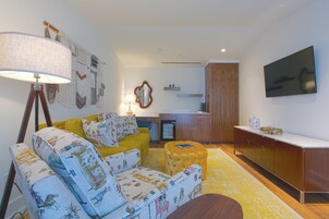 Un salón con sofá, reposapiés y lámpara. Hay un televisor montado en la pared y una pequeña zona de cocina con un refrigerador y un microondas.