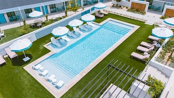 Una piscina con sillas de descanso y sombrillas, rodeada de un césped cuidado y una fachada de edificio moderna.