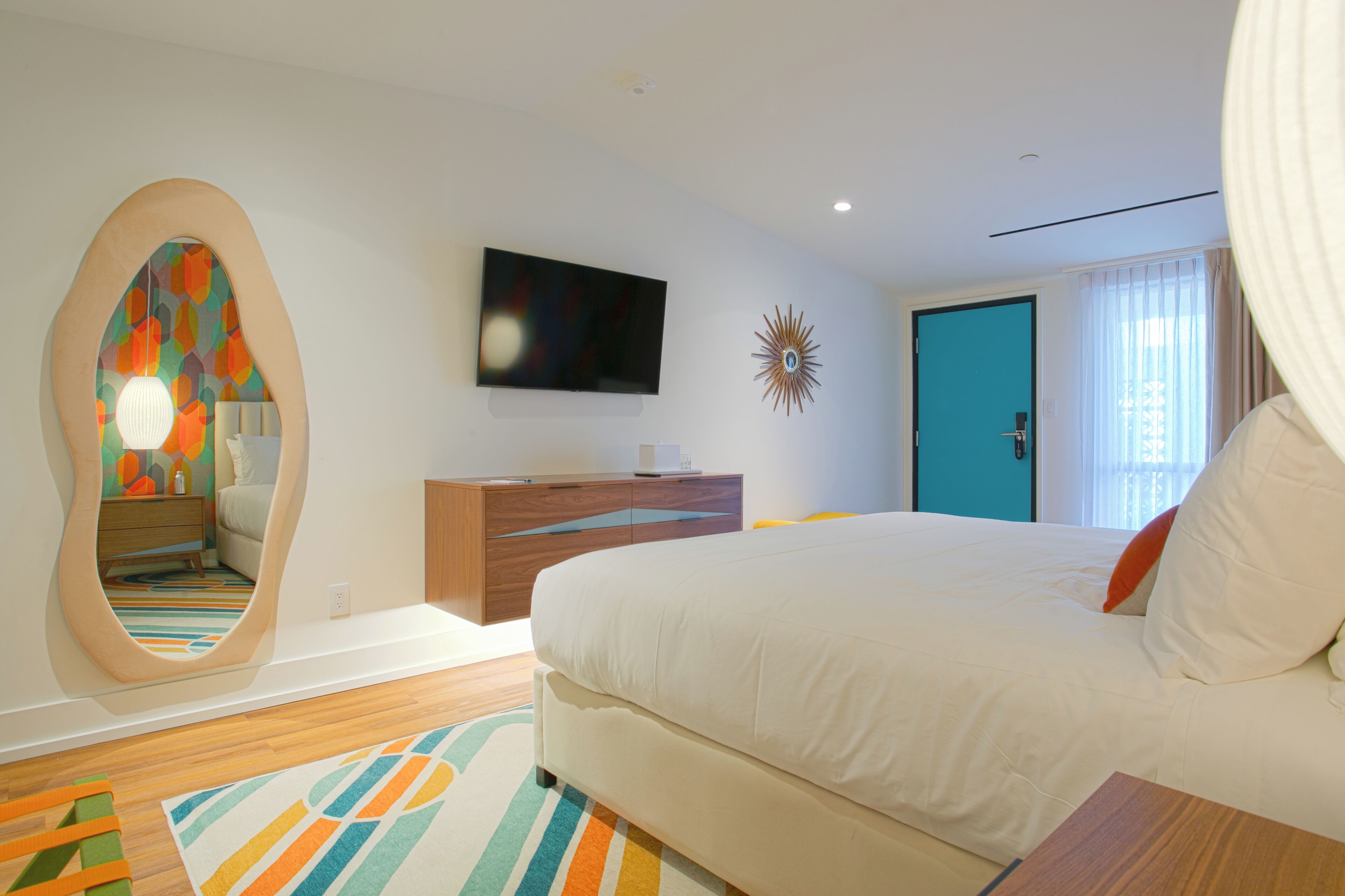 A modern hotel room with a large bed, a TV mounted on the wall, a mirror with a colorful pattern, and a wooden dresser.