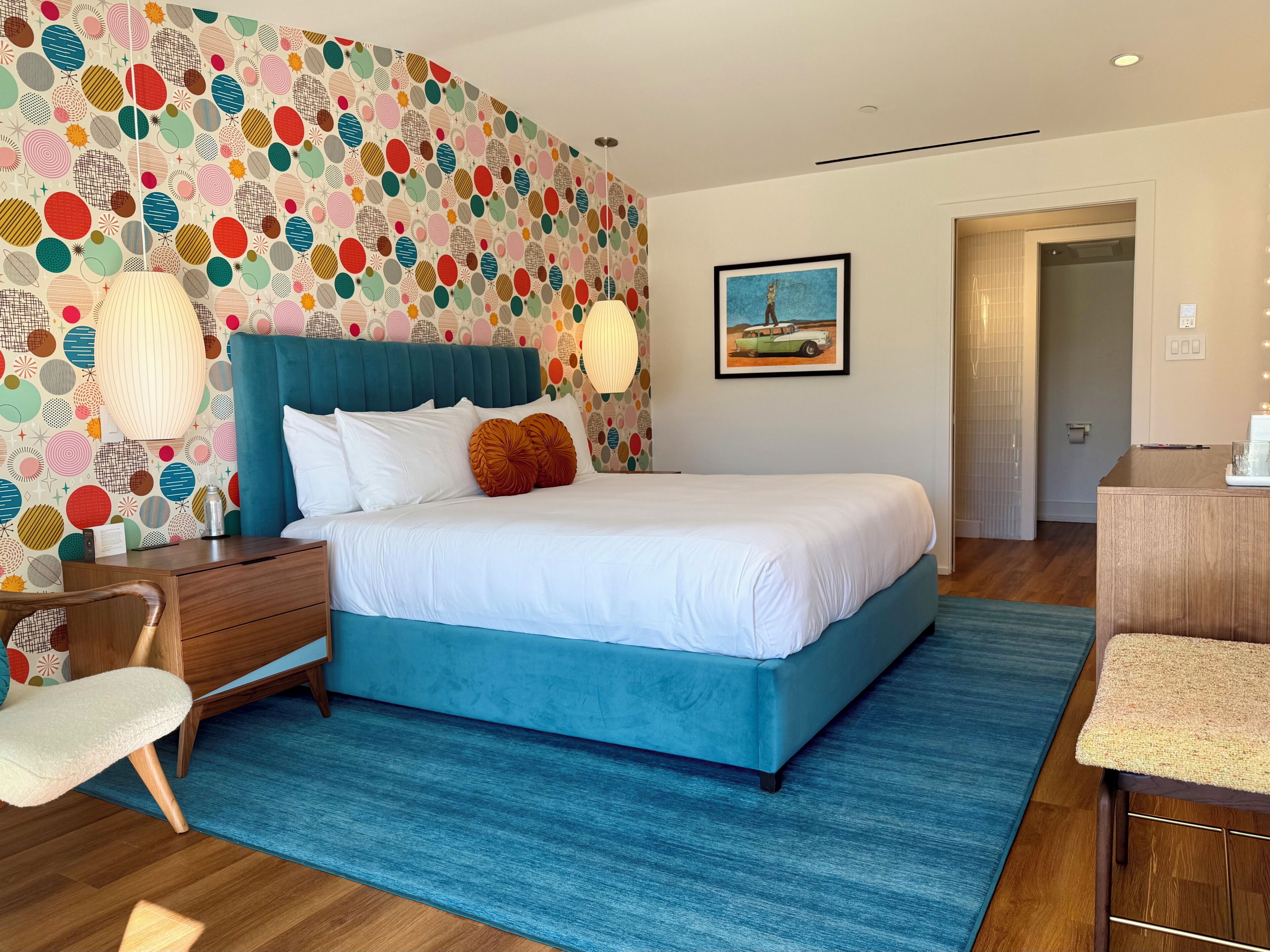 A modern bedroom with a teal bed, white bedding, and a colorful circular-patterned wallpaper.