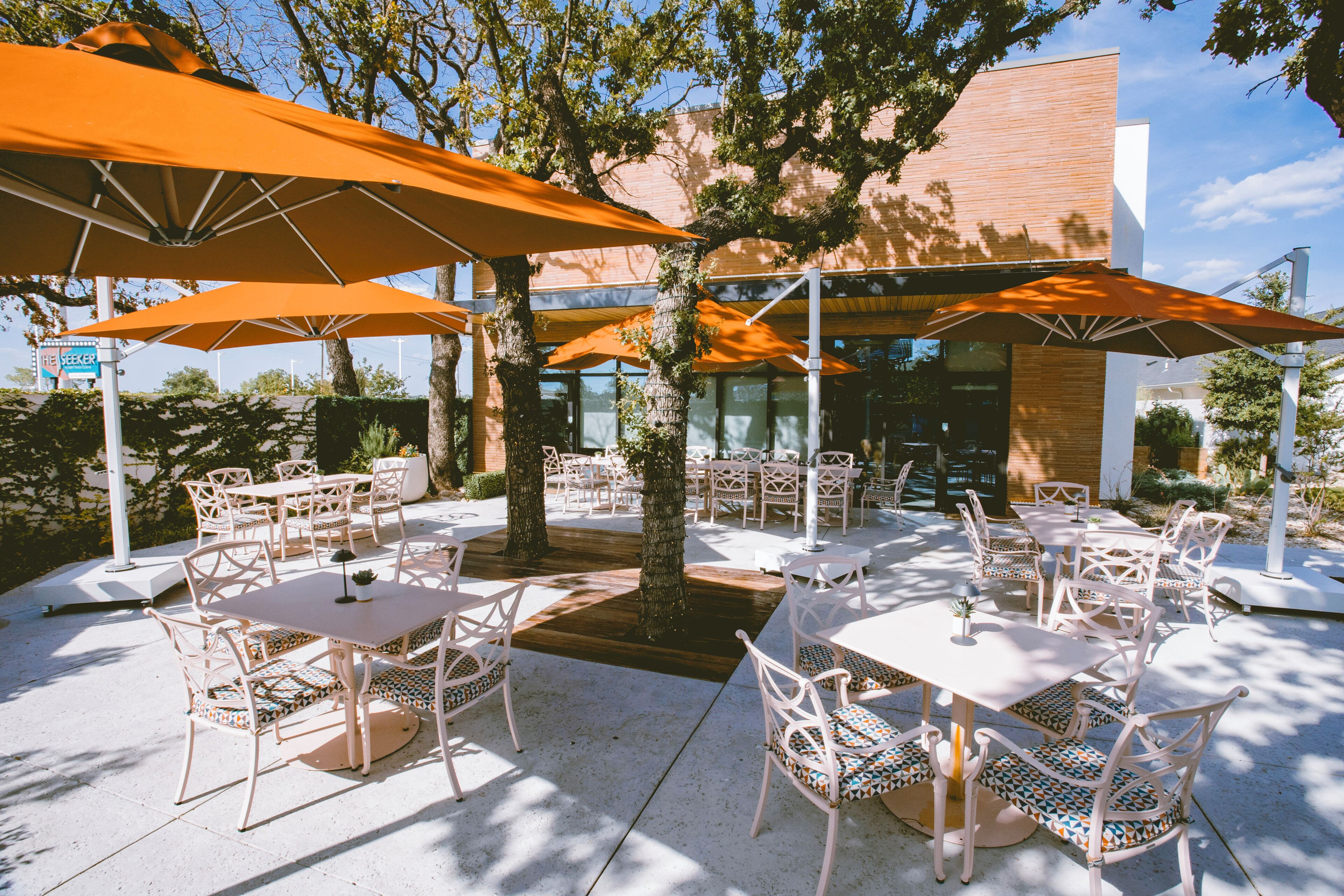An outdoor seating area with tables and chairs, surrounded by trees and covered by orange umbrellas.