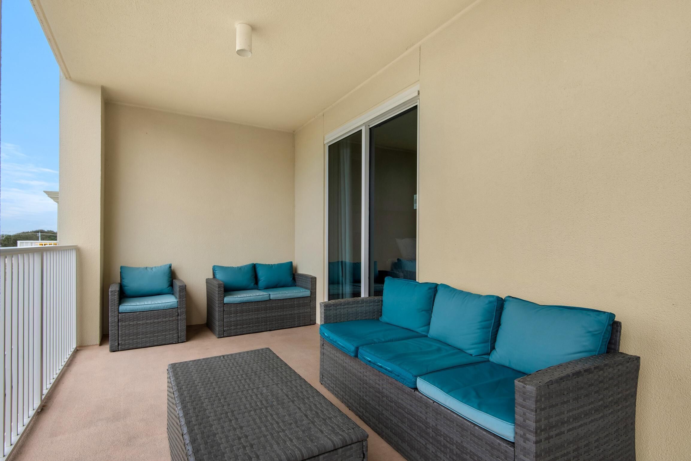 A balcony with two grey wicker sofas and a matching ottoman, each with teal cushions.