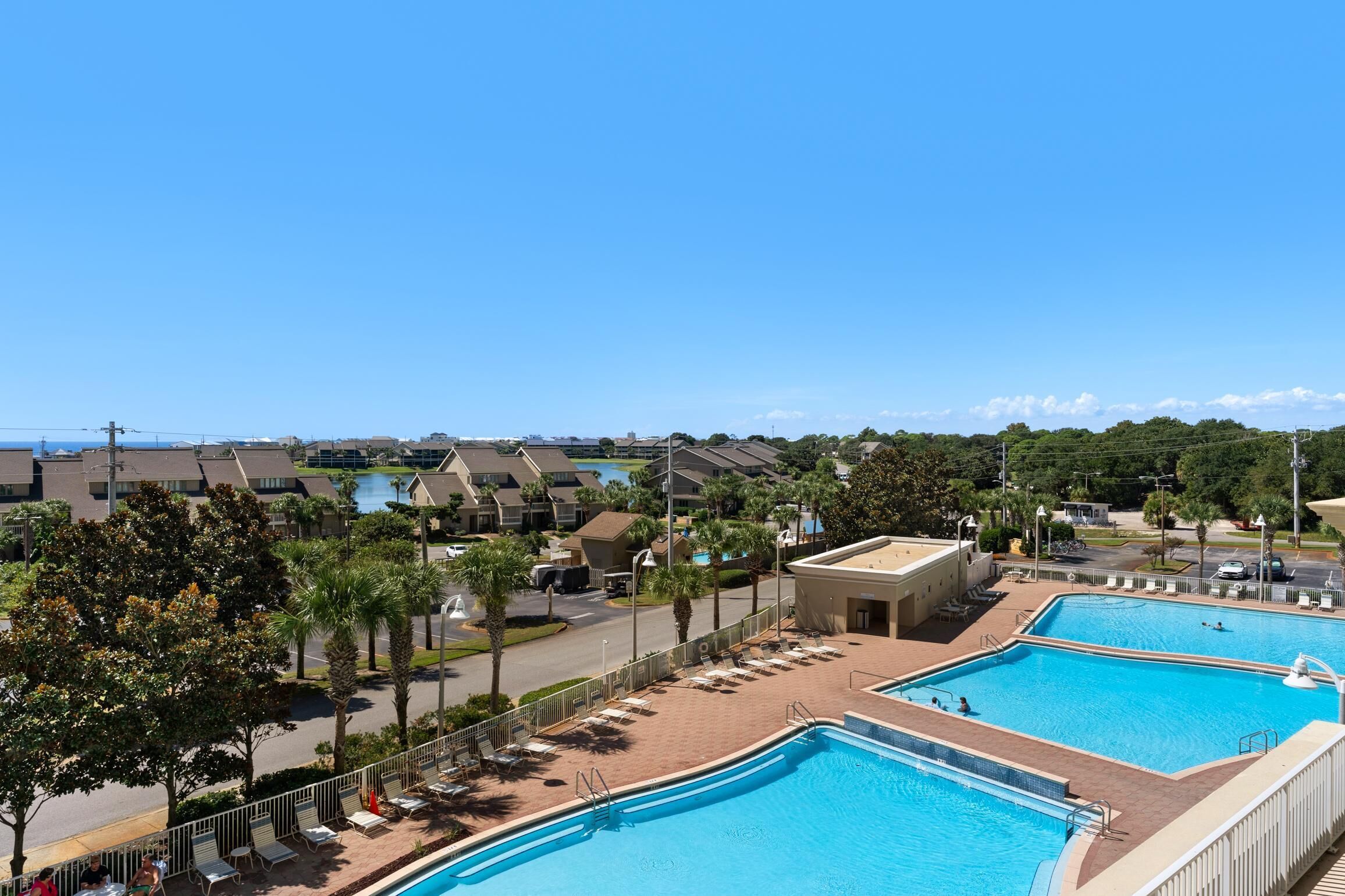A resort with two swimming pools, a road with parked cars, and a view of a body of water and buildings in the distance.