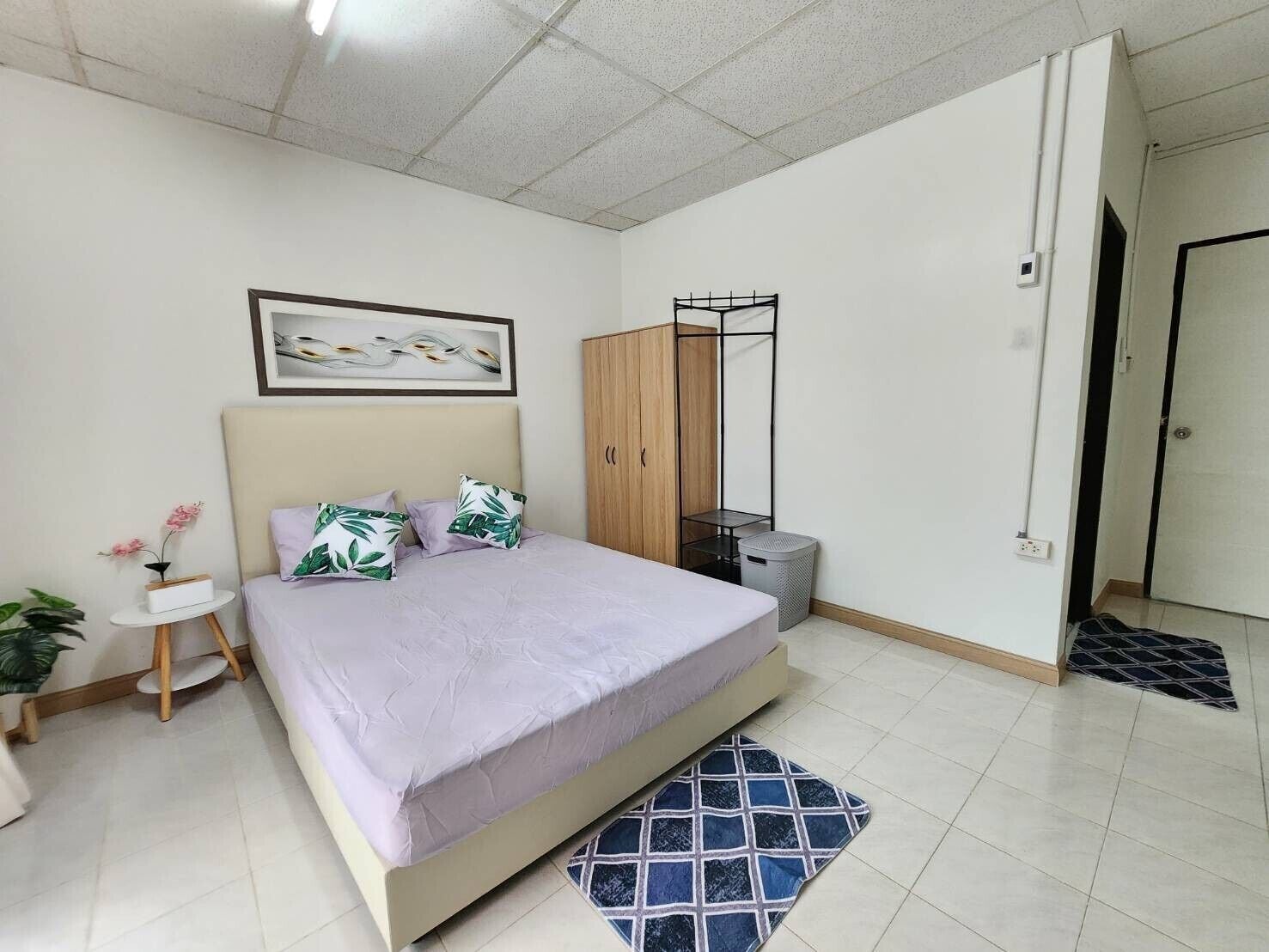 A bedroom with a bed, a wardrobe, a small table with a plant, and a trash bin.