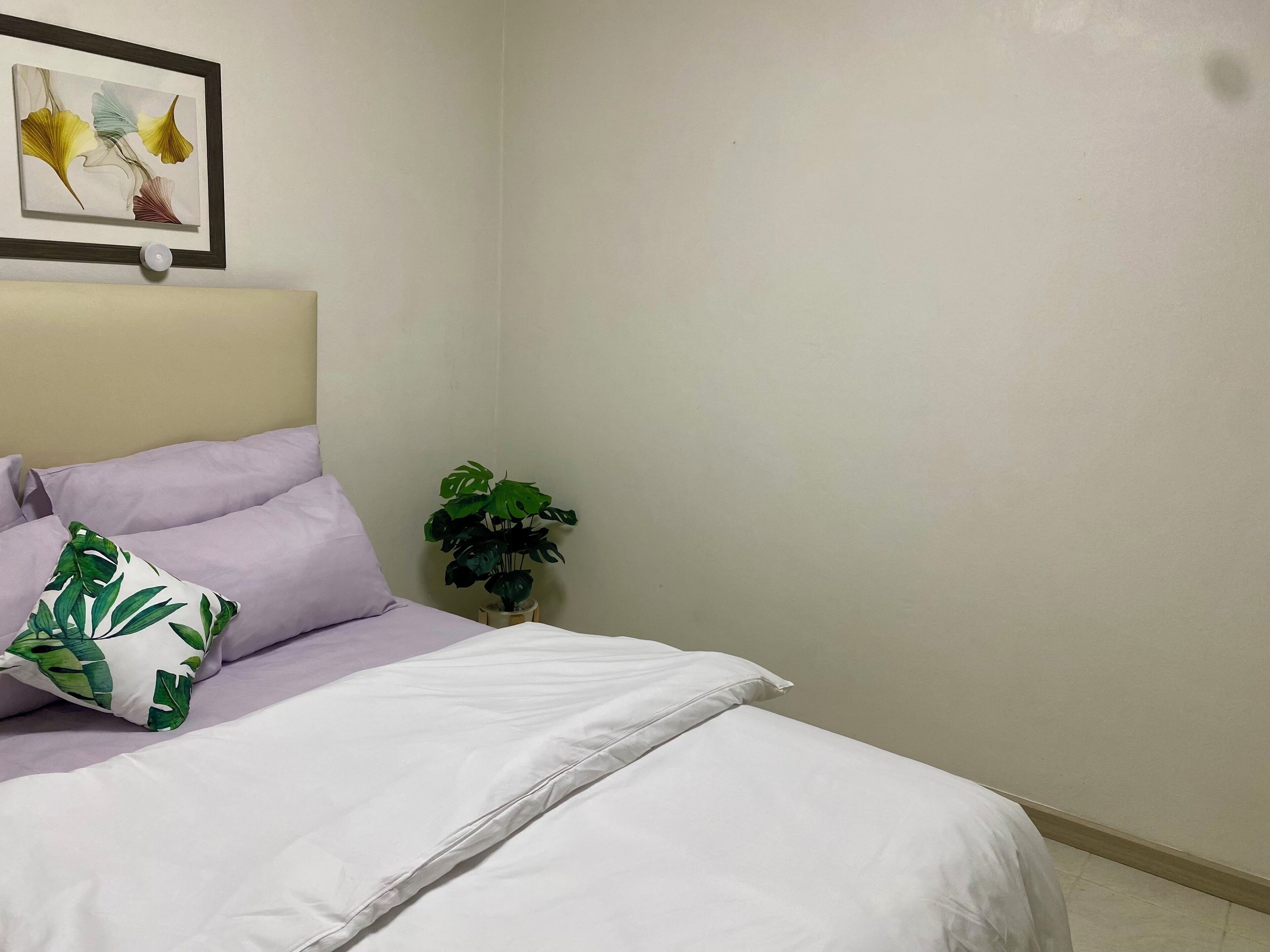 A neatly made bed with white and lavender bedding, a framed artwork above it, and a potted plant beside the bed.