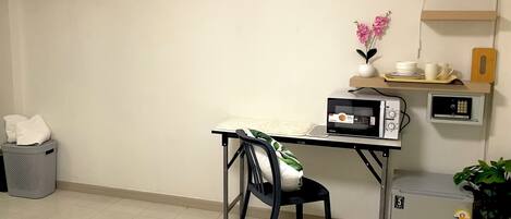 A compact hotel room with a small desk, a microwave, a refrigerator, a chair, a trash bin, a fan, and a potted plant.