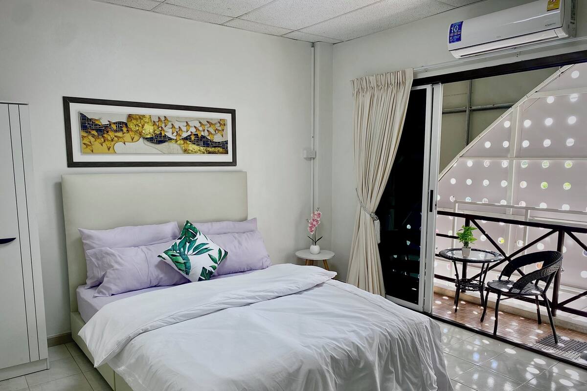 Un dormitorio moderno con cama, una obra de arte enmarcada encima, una mesita de noche con un florero y un balcón con mesa y sillas.