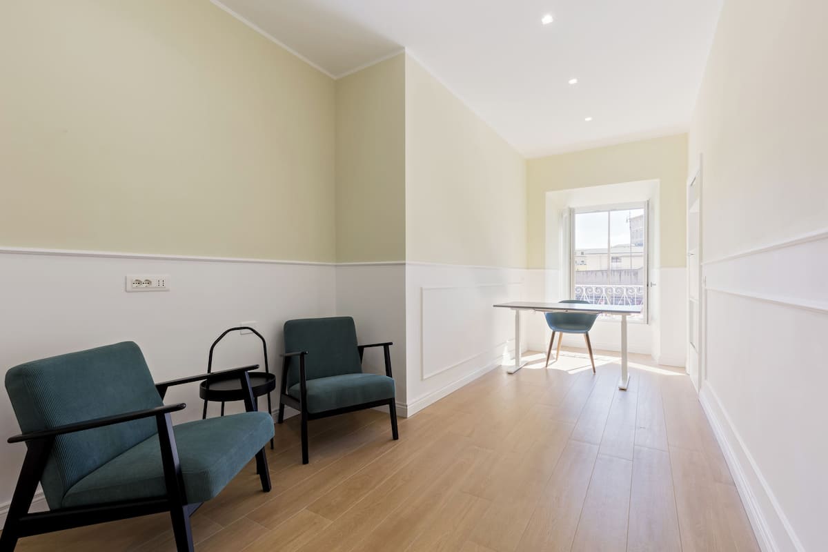 A room with two teal chairs, a small table, and a window with a view of buildings.