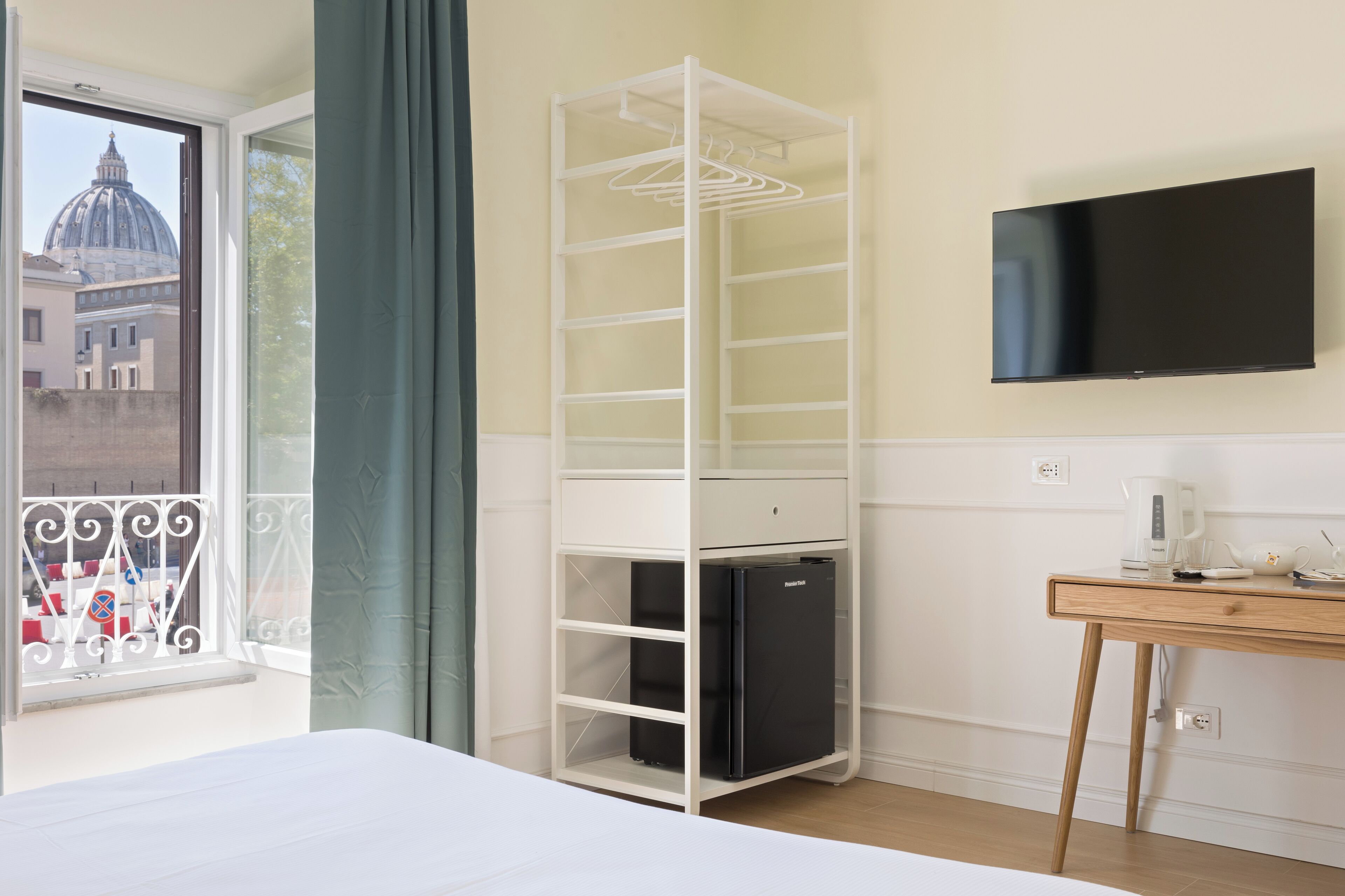 A hotel room with a bed, a white shelving unit with a black refrigerator, a wooden table with a coffee maker, a TV on the wall, and a view of a building with a dome through a window.