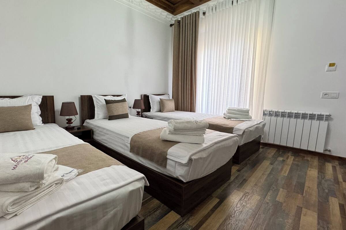 A hotel room with two single beds, a large window with curtains, a radiator, and a wooden floor.