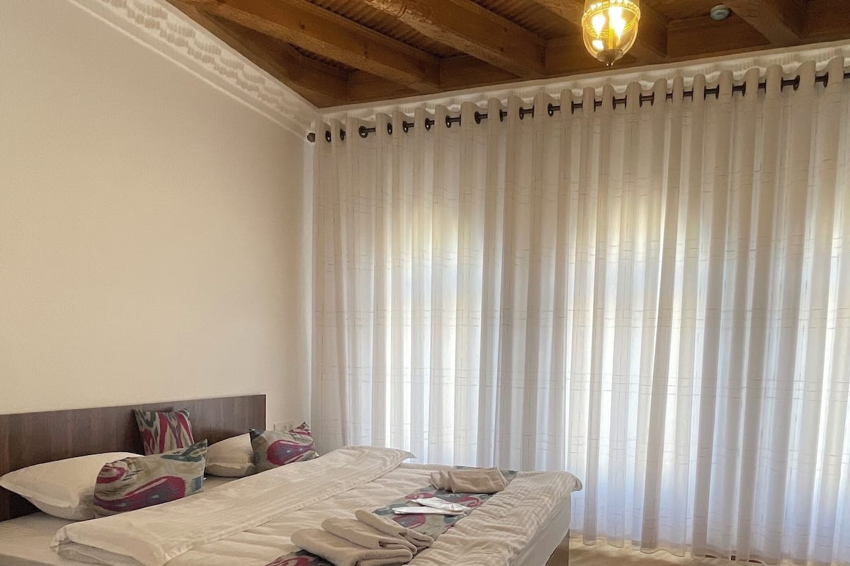 A bedroom with a wooden ceiling, a bed with patterned bedding, and a large window with sheer curtains.