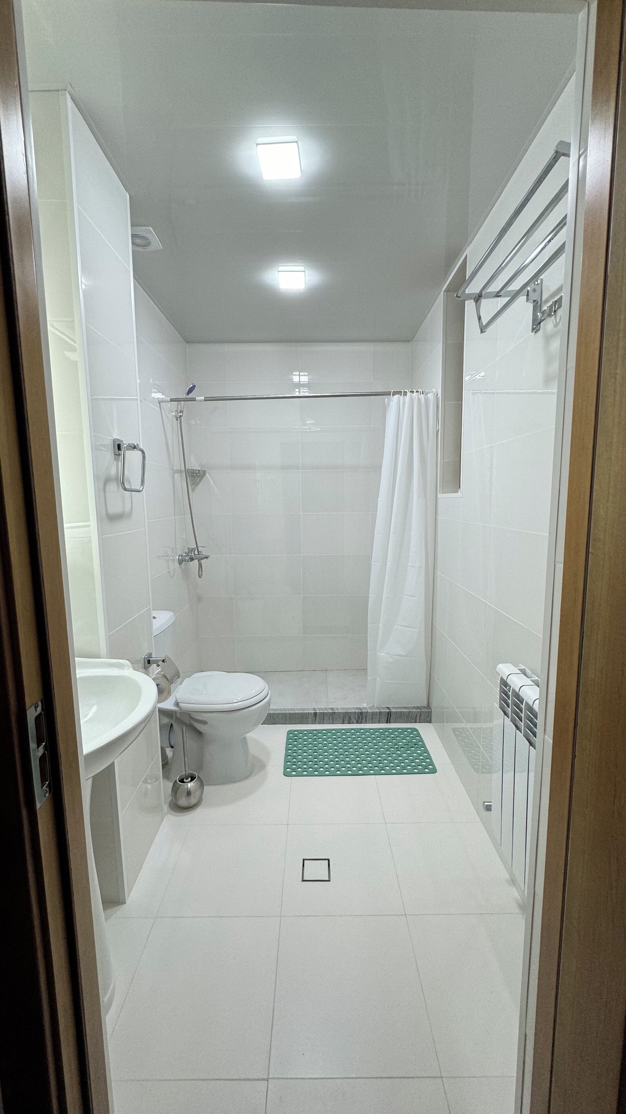 A modern bathroom with a glass shower enclosure, white tiles, a toilet, a sink, and a green mat.