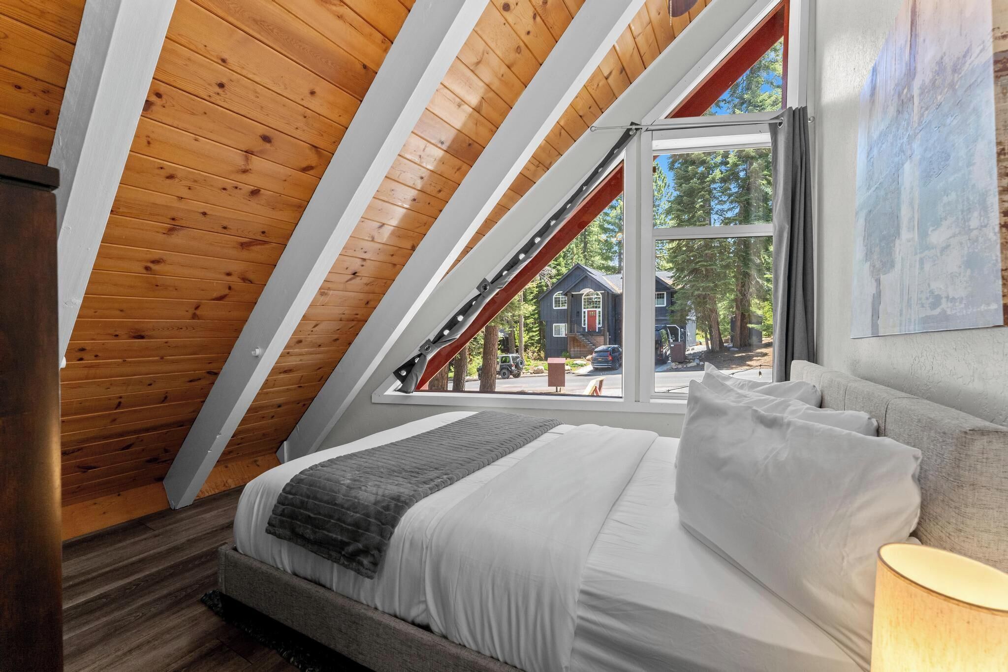 Un dormitorio con techo inclinado, cama con ropa de cama blanca, una manta gris y una ventana con vista a una casa y árboles.