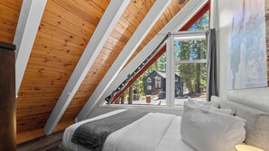 A bedroom with a sloped ceiling, a bed with white bedding, a gray throw blanket, and a window with a view of a house and trees.
