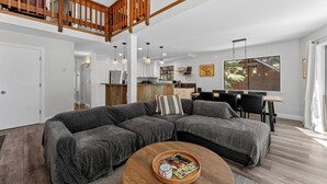 A modern living room with a sectional sofa, a coffee table, and a dining area with a wooden table and chairs. A kitchen is visible in the background.