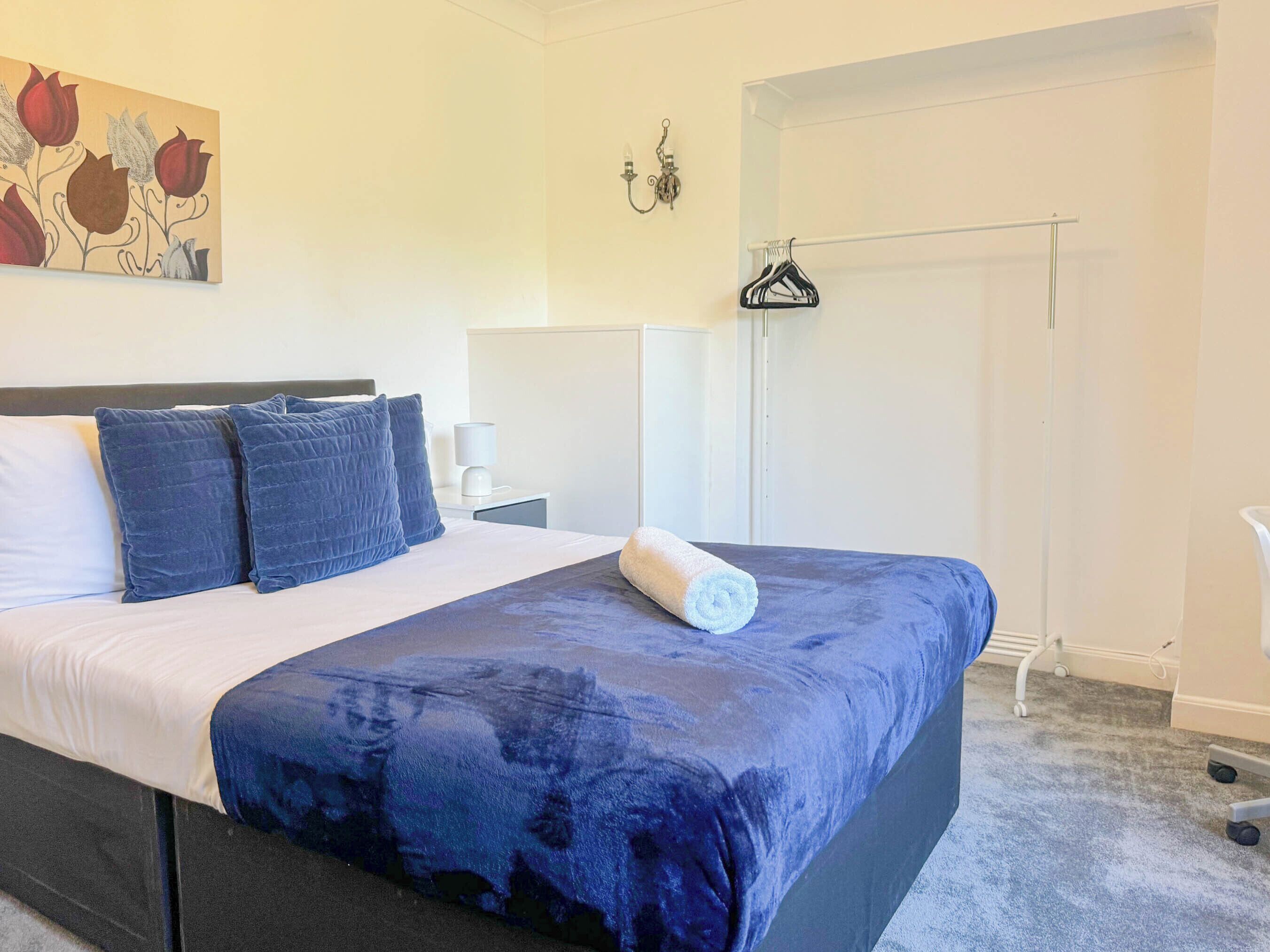 Ein Schlafzimmer mit einem Bett, Kissen, einem Handtuch, einer Wandleuchte, einem Kleiderhaken und einem Gemälde.