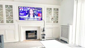 A living room with a white fireplace, a flat-screen TV mounted above it, and a white entertainment unit with glass doors. - Large 8 Bedroom House in Edgbaston (Birmingham)