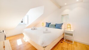 Une chambre moderne avec un plafond en pente, un grand lit recouvert de draps blancs, des oreillers bleus et une table de chevet avec une lampe.