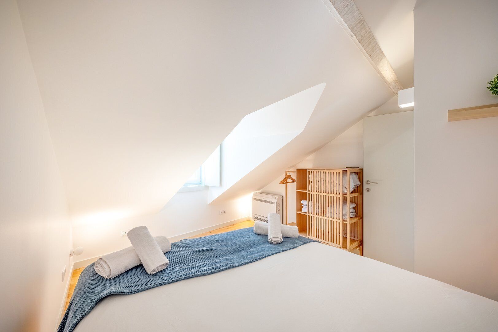 Ein modernes Schlafzimmer mit schrägem Dach, einem Bett mit aufgerollten Handtüchern, einem Holzschrank und einer kleinen Pflanze.