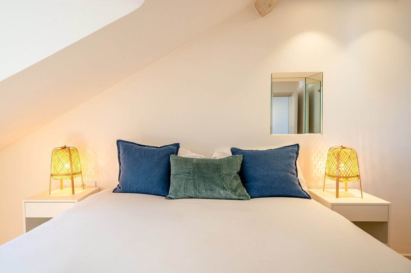 A bedroom with a sloped ceiling, a bed with white bedding, two blue pillows, a green pillow, and two bedside lamps with yellow shades.
