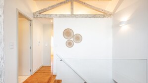 Un escalier moderne avec des marches en bois et une rampe en verre, menant à une pièce aux murs blancs et dont le plafond est orné de poutres apparentes.