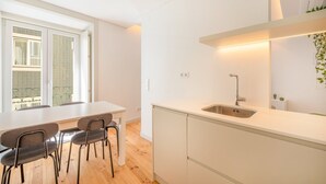 Una cocina moderna con encimera blanca, un lavaplatos de acero inoxidable y una zona de comedor con mesa y sillas.