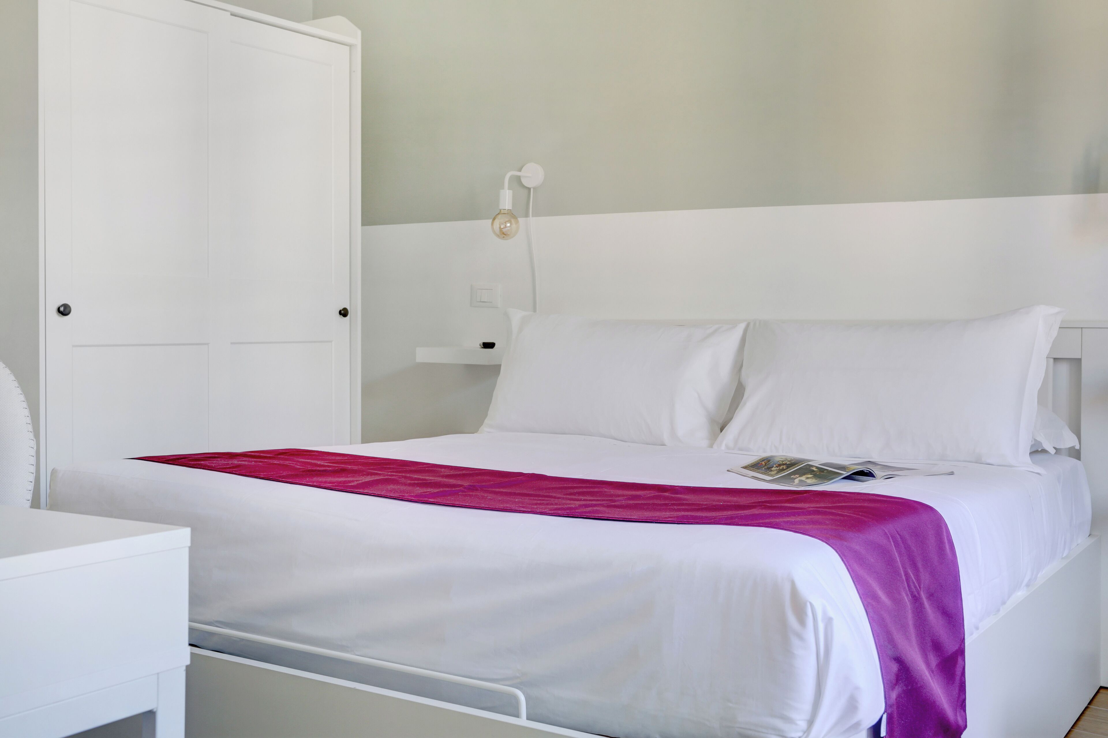 A neatly made bed with white bedding and a pink accent, a white wardrobe, and a bedside table with a lamp.