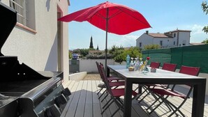 Patio esterno con barbecue, tavolo e sedie, e un ombrellone rosso.