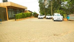 A paved driveway leading to a building with a modern design, two white cars parked in front, and a small garden area with a tree and a planter. - Hirwa Avenir (Kigali)