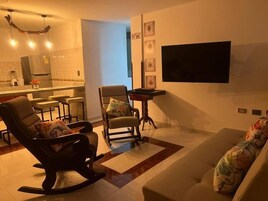 Una sala de estar con un sofá, un sillón mecedora, una mesita y un televisor montado en la pared.