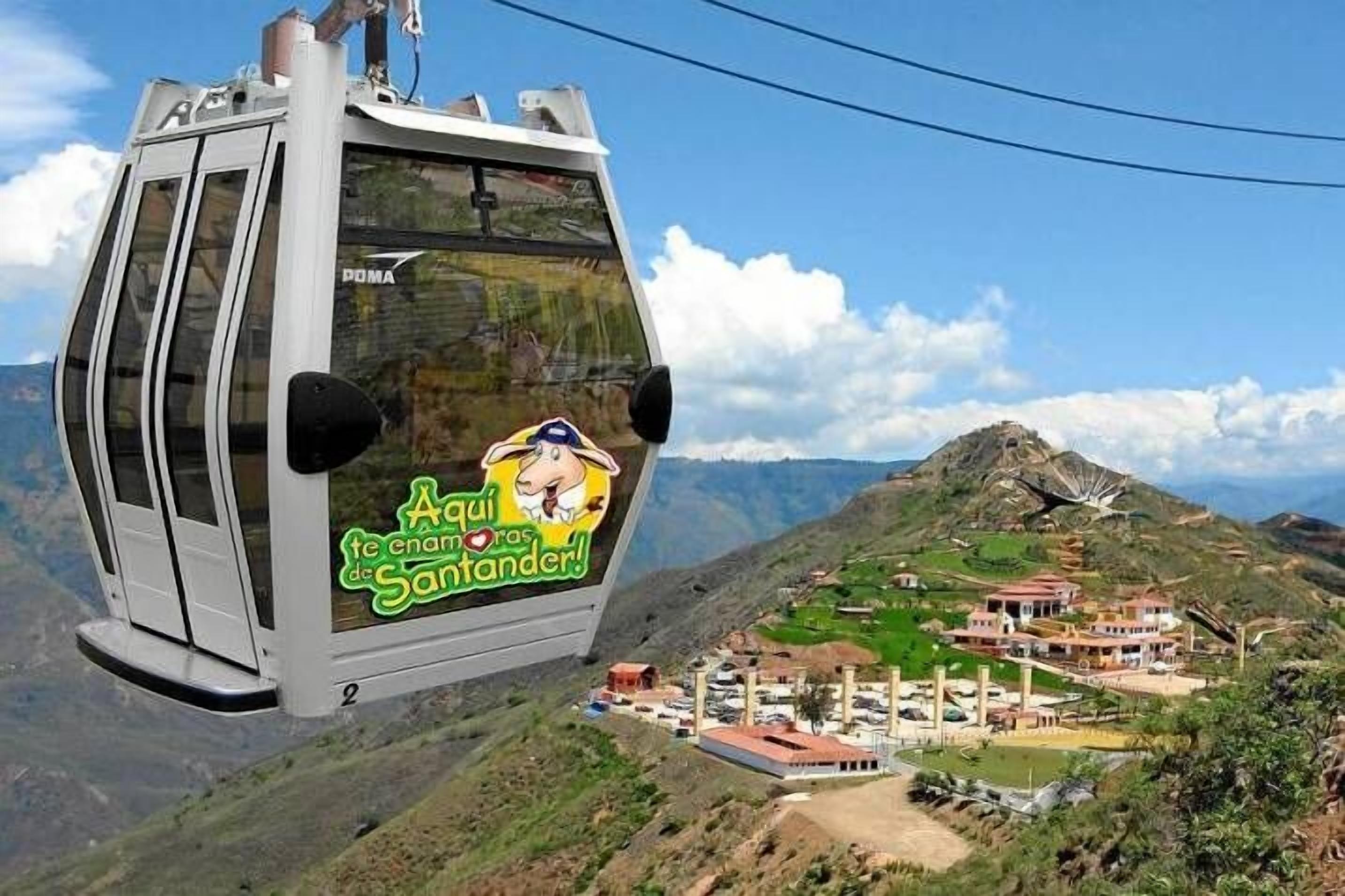 A cable car with a "Aqui enamor de Santander!" sign, flying over a mountainous landscape with buildings below.