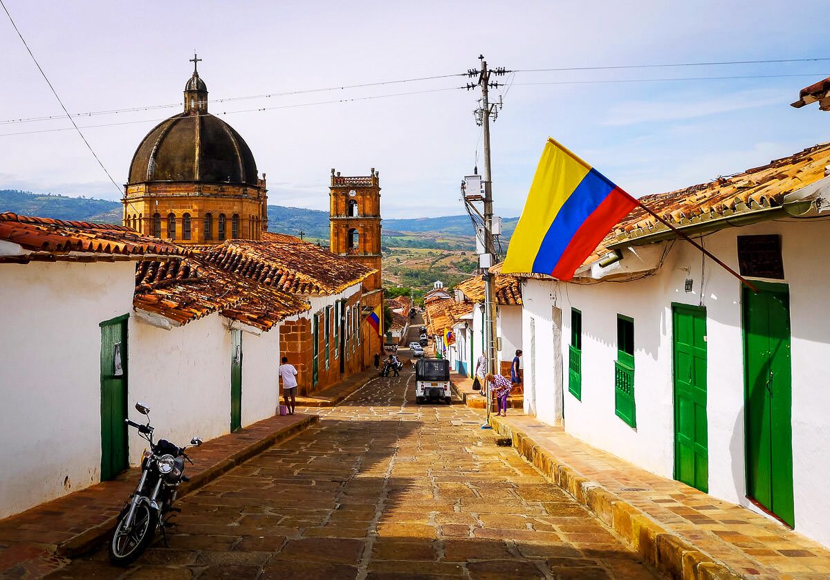 A narrow cobblestone street with historic buildings, a church with a dome, and a flag.