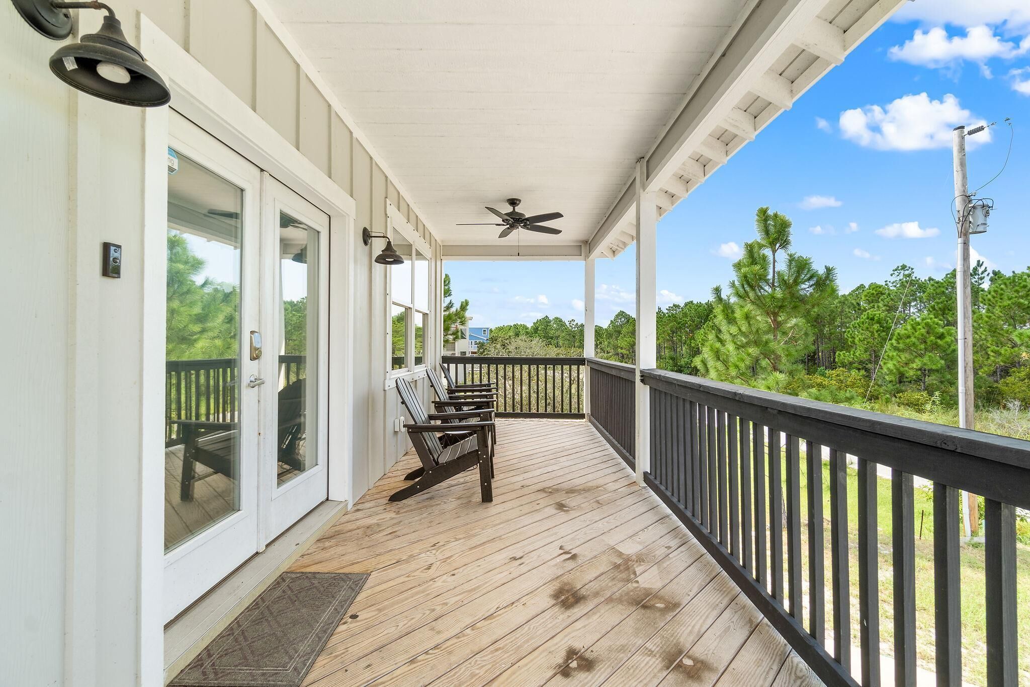 A covered porch with a ceiling fan, wooden deck, and a view of greenery.