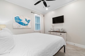 A bedroom with a bed, a ceiling fan, a wall-mounted TV, a window with blinds, and a decorative piece on the dresser. - Beach Please (Gulf Shores)