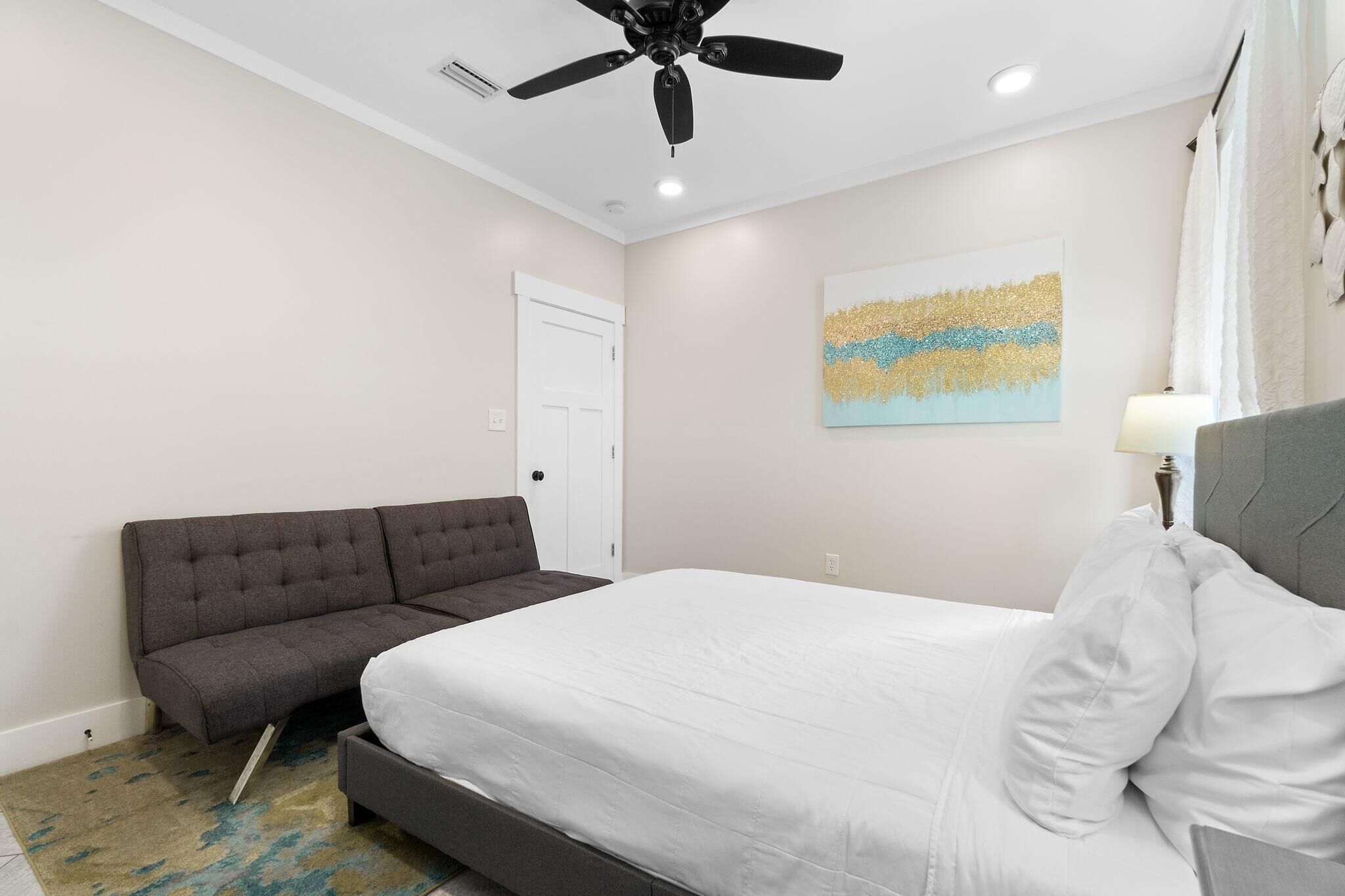 A bedroom with a bed, a sofa, a ceiling fan, a painting, and a lamp.