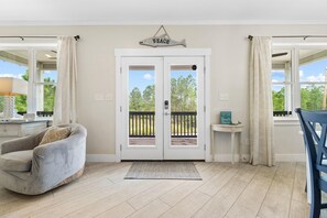 Una habitación con una puerta doble que da a un balcón, un sillón gris, una mesita con un cartel y vistas a árboles y cielo.
