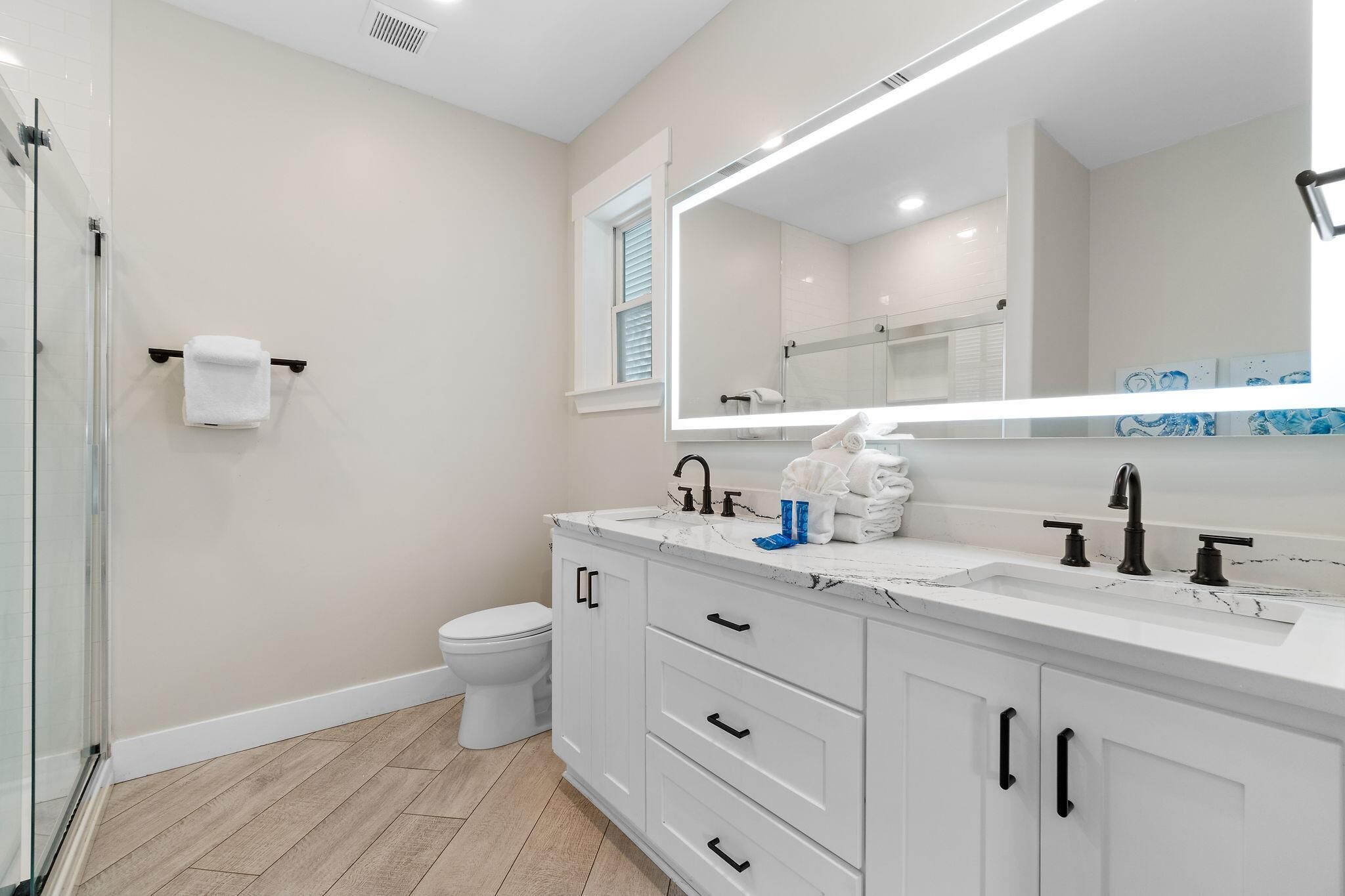 A modern bathroom with a glass shower enclosure, a white toilet, a large mirror with integrated lighting, and a white vanity with black handles.