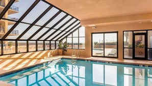 Une piscine intérieure avec des parois en verre transparent, offrant une vue sur l’extérieur, et un espace aménagé au bord de la piscine avec des chaises et un parasol.