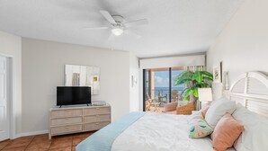 Une chambre à coucher avec un lit, un téléviseur posé sur une commode, un ventilateur de plafond et un balcon offrant une vue sur l’océan.