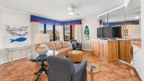 Un salon comprenant un canapé, un fauteuil, une table basse et un téléviseur. On y trouve également un ventilateur de plafond, une fenêtre avec des rideaux et un objet décoratif en forme de planche de surf.