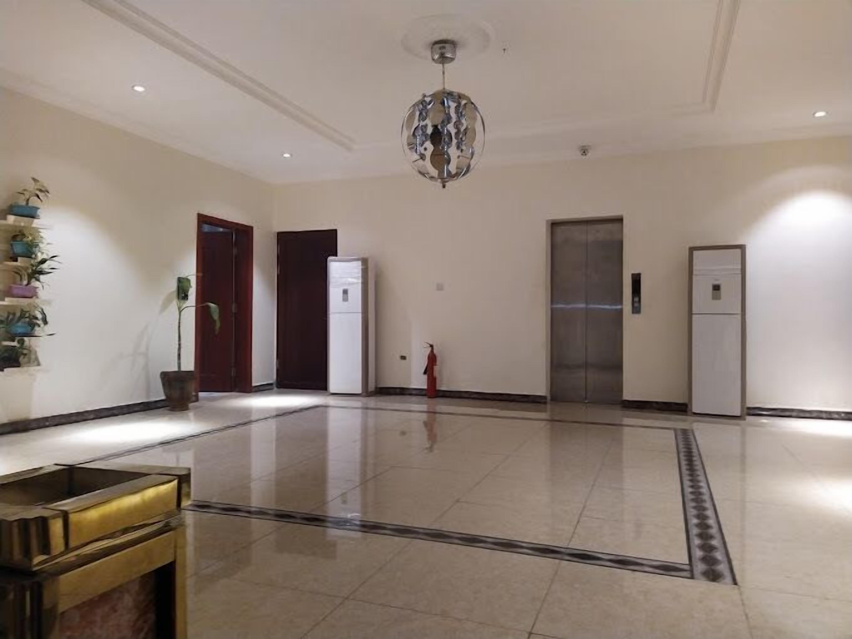 A spacious lobby with a polished floor, a decorative chandelier, a refrigerator, an elevator, and a fire extinguisher.