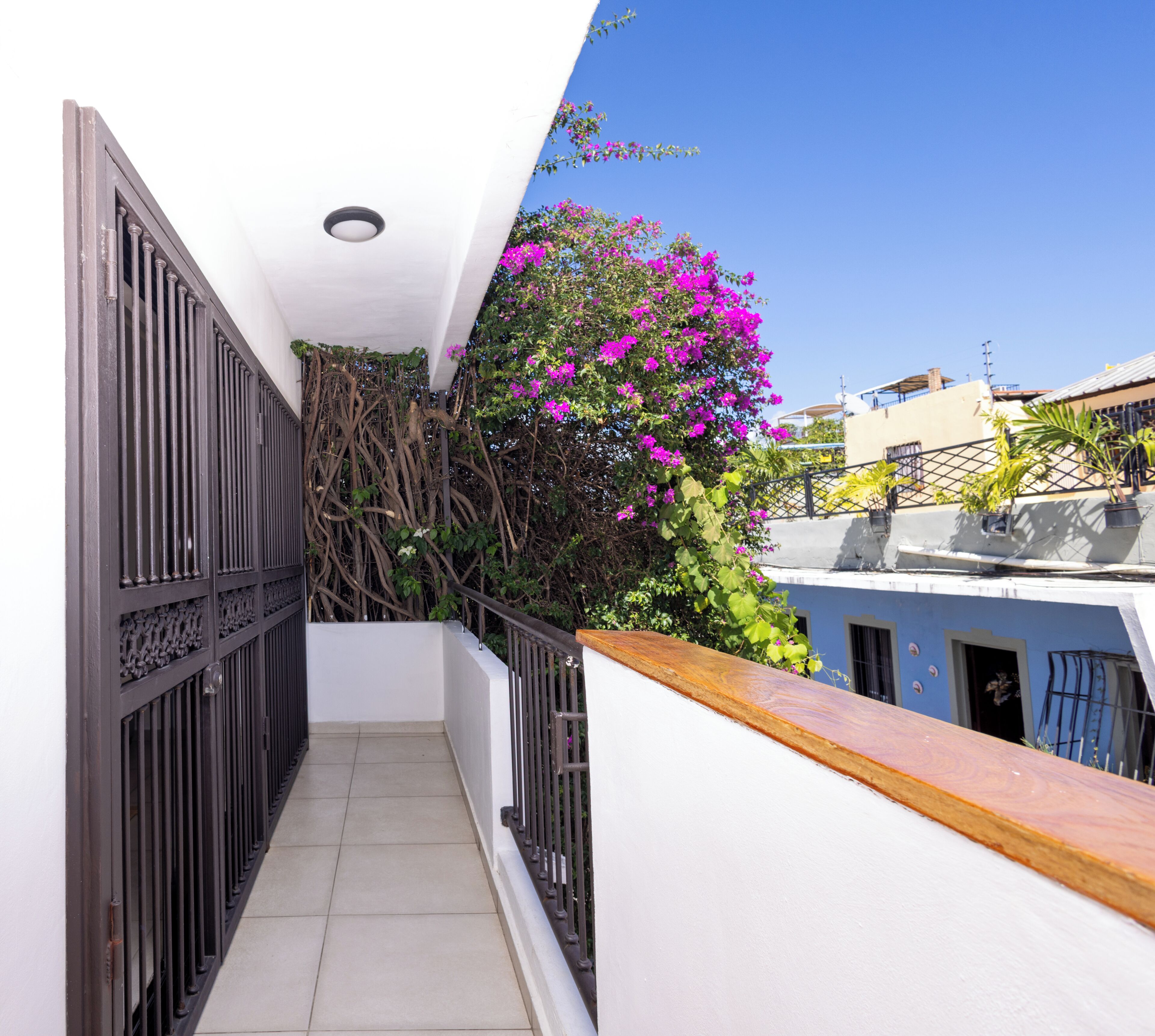 Un balcon étroit doté d’un sol carrelé, d’une balustrade en bois et offrant une vue sur un immeuble à la façade bleue et aux fleurs roses.