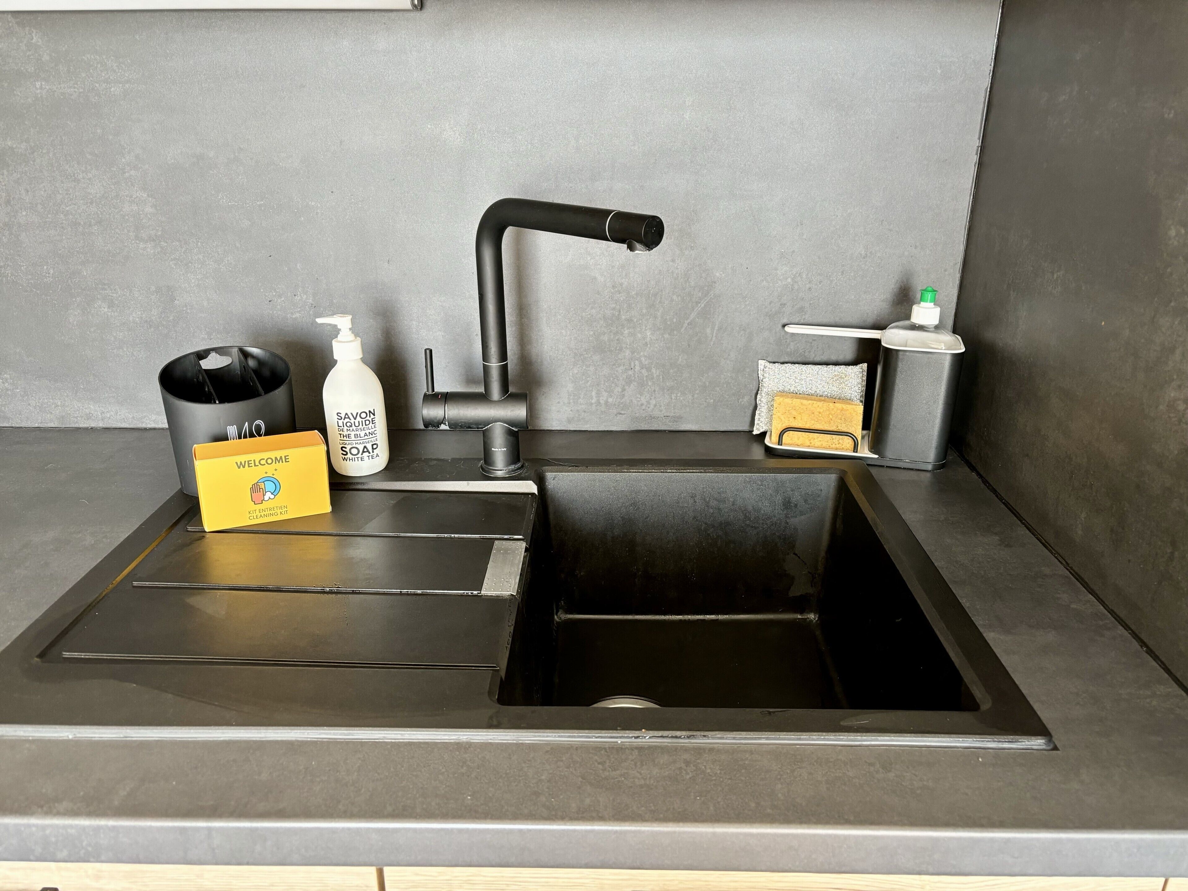 A modern kitchen with a black sink, a soap dispenser, a sponge, and a welcome card.