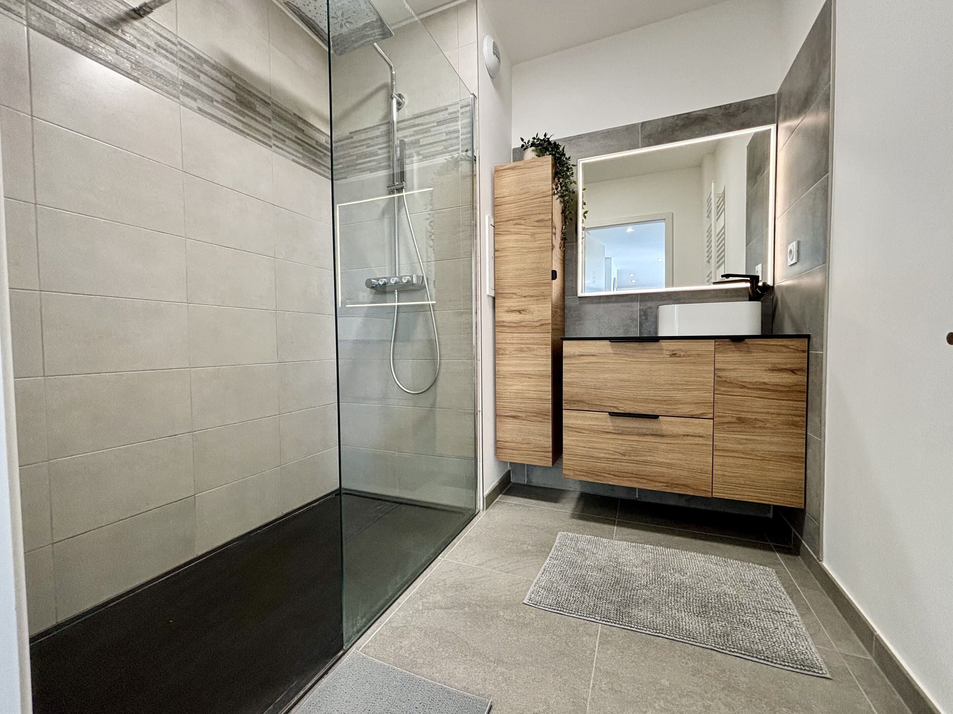 Une salle de bain moderne avec une douche à parois de verre, un meuble-lavabo en bois avec un miroir et un sol carrelé.