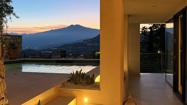 A modern outdoor terrace with a pool, plants, and a view of a mountain range at dusk. - Villa Ada: Elegant Villa with Private Pool & Mount Etna Views (Taormina)