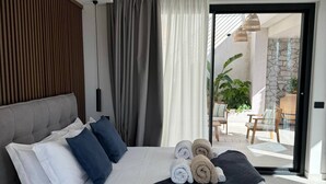 A modern bedroom with a large bed, a wooden headboard, and a view of an outdoor patio with chairs and a plant. - Villa Ada: Elegant Villa with Private Pool & Mount Etna Views (Taormina)