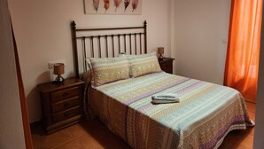 A bedroom with a wooden bed, a nightstand with a lamp, a dresser, and a wall-mounted artwork.